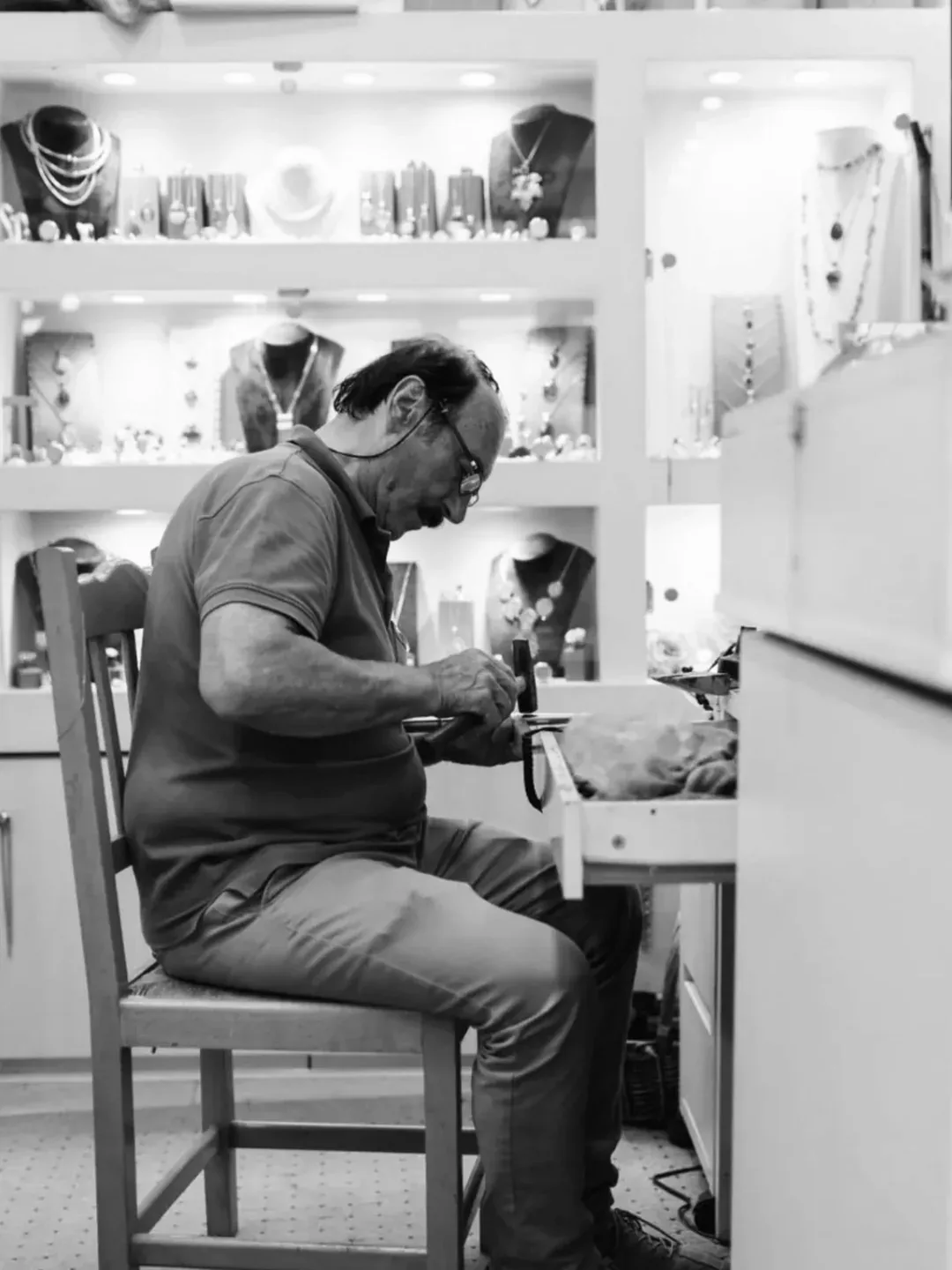 Ασπρόμαυρη φωτογραφία ενός χρυσοχόου σε προφίλ, καθισμένου σε ξύλινη καρέκλα και σκυμμένου πάνω στον πάγκο του, που σφυρηλατεί ένα κομμάτι μετάλλου, με φωτισμένες βιτρίνες με κοσμήματα σε προτομές πίσω του.